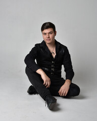 Full length portrait of a  brunette man wearing black shirt and gothic waistcoat.  Seated  pose...
