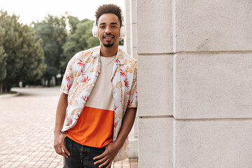 Brown-eyed curly brunet young man in colorful t-shirt and floral shirt smiles sincerely, leans on...