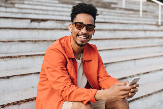 Joyful dark-skinned curly man in orange jacket and sunglasses smiles widely, holds phone and sits on stairs outside.