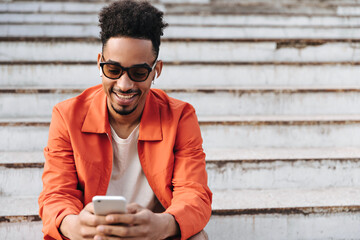 Excited young charming bearded man in sunglasses and orange jacket smiles sincerely, sits on stairs and holds phone outside.