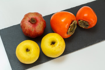 Apples, pomegranate and ripe persimmon fruit on the black stone board.