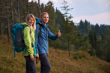 Two young tourists with joyful facial expressions holding hands while walking outdoors. Man pleased with hike at mountain trails and lawns showing thumb up. Concept of travelling and hiking.