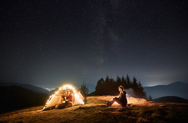 Young man traveler sitting inside illuminated camp tent and playing guitar while spending time with girlfriend in mountains under blue night sky with stars. Concept of night camping and relationships.