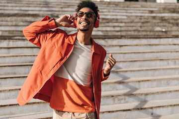 Excited brunet charming man in orange jacket, colorful t-shirt and sunglasses sings, sincerely...
