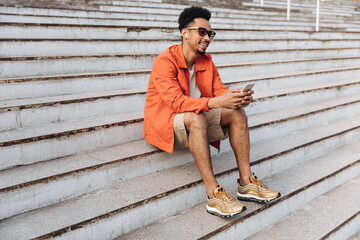 Cool young curly dark-skinned man in orange jacket and beige shorts sits on stairs outside. Happy...