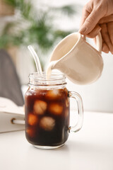 Woman pouring milk from jug into mason jar with tasty ice coffee on table, closeup