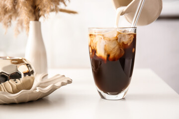 Pouring milk from jug into glass with tasty ice coffee on table, closeup