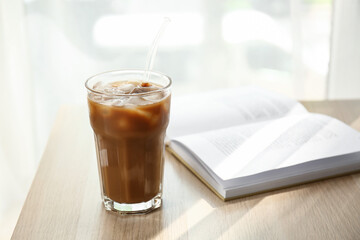Glass of tasty ice coffee with milk and book on wooden table