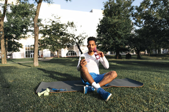 Calm handsome man in blue shorts and white t-shirt sits on yoga mat in park, holds phone and drinks water.
