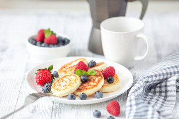 Cottage cheese pancakes with fresh berries, cup of coffee and coffee maker on white wooden table. Tasty breakfast food. Syrniki