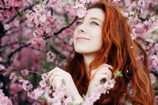 Stylish russian beauty with long red loose hair smiling and looking into the distance. Sensual portrait of a girl posing against a background of blurred pink flowers