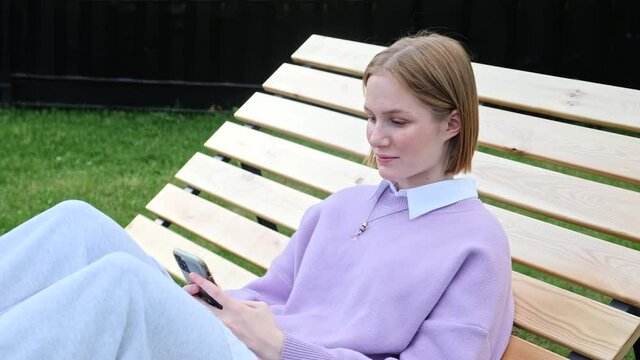 Young woman student looking at smartphone screen watching funny photo or video, enjoying online communication in social network web surfing or reading good comments and smiling. Outside in the park