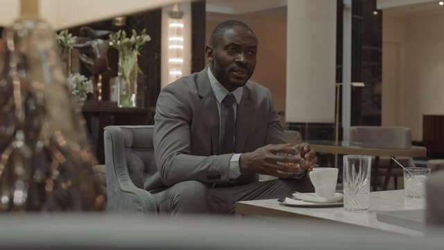 Medium long of young African professional man wearing grey formal suit, sitting in armchair in hotel lobby, holding rocks glass, talking to invisible person across table