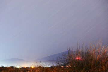 Gloomy spring background with star trails and lights of a distant city next to low mountains....