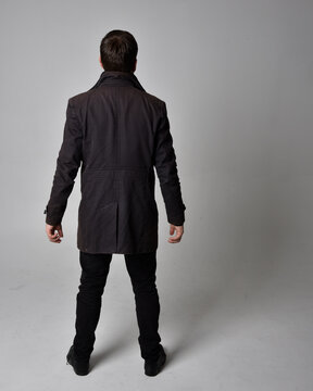 Full Length Portrait Of A Brunette Man Wearing Black Leather Coat.  Standing Pose Facing Backwards, Walking Away From The Camera Isolated  Against A Grey Studio Background.