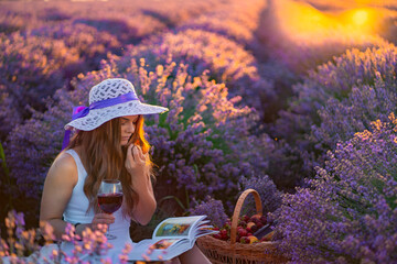 young beautiful attractive lady in white dress with white hat reads a book and holds a glass of red wine in lavender field during sunrise
