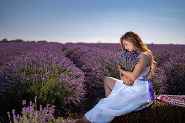 beautiful tender young lady holding a bouquet of lavender, sitting on a haystack with lavender field on background © Sid Smith