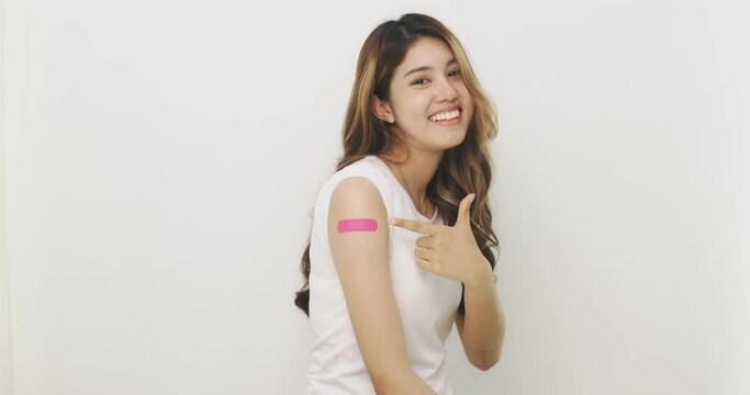 Asian Woman Points To Bandage On Arm. Happy Asian Woman Feels Good After Received Vaccine On White Background.