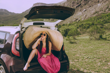 Young beautiful woman in a dress and a wide-brimmed hat near a hatchback car on a background of a mountain landscape.