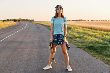 Full length photo of confident satisfied female wearing short, t shirt, hair band, holding skateboard in hands, looking at camera, after skateboarding outdoor.