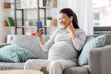 Obraz premium pregnancy, rest, people and expectation concept - happy smiling pregnant asian woman with smartphone and earphones sitting on sofa at home and listening to music