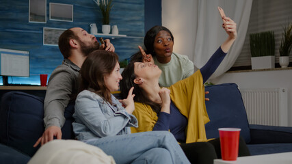 Fototapeta premium Excited group of multiracial friends sitting on sofa during joyful party taking selfie posting on social media. Multi-ethnic friends enjoying time together sharing photos late at night