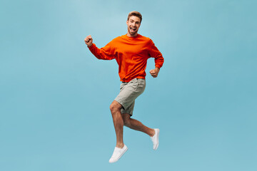 Fototapeta na wymiar Full length photo of emotional young guy in orange sweatshirt, beige shorts and white sneakers jumping and smiling on isolated backdrop..