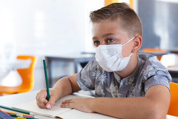 Caucasian schoolboy wearing face mask sitting at desk in classroom writing in book