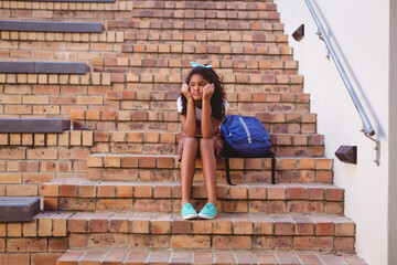 Bored african american schoolgirl sitting on steps in school yard with schoolbag