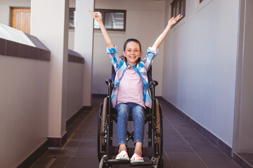 Portrait of smiling disabled caucasian schoolgirl sitting in wheelchair raising arms in corridor
