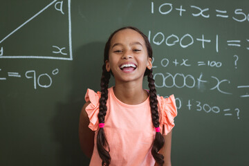 Portrait of laughing mixed race schoolgirl standing in front of chalkboard in classroom maths lesson - Powered by Adobe