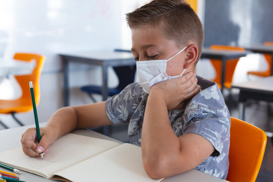 Caucasian schoolboy wearing face mask sitting at desk in classroom writing in book