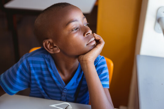 Bored african american schoolboy sitting at desk looking out of window in classroom