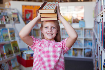 Portrait of caucasian schoolgirl smiling carrying stack of books on head in school library