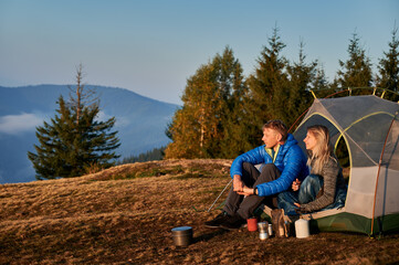 Naklejka na ściany i meble Side view of married couple of travelers resting in tent on mountain meadow during autumn hike against the backdrop of trees and mountain hill. Sunshine morning in campsite.