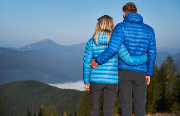 Rear view of couple hikers holding each other around waist and contemplating on fabulous mountain landscape in front of them at sunshine autumn vacation.