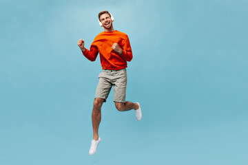 Fototapeta premium Joyful fashionable guy with red beard in bright orange cool clothes listening to music and jumping on blue isolated backdrop..