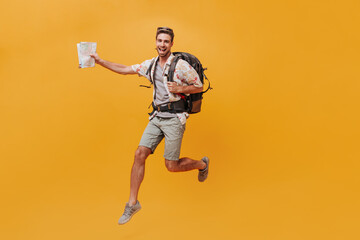Fototapeta na wymiar Slim happy man with beard in white summer shirt and grey shorts jumping with large backpack and map on isolated background..