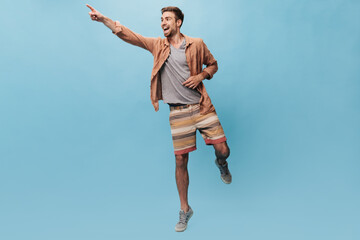 Fototapeta premium Full length photo of cheerful man with beard in cool light t-shirt and striped unusual shorts looking away and showing aside his finger..