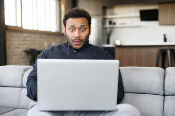 Shocked indian young man staring at laptop display, feel astonishment with a bad news, fired from work, irritated eastern guy looks at computer screen and does not understand what happened