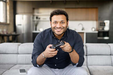 Handsome excited young indian man spends leisure time playing video game, hispanic guy with a joystick sitting on the couch, play on console at home, front view © Vadim Pastuh