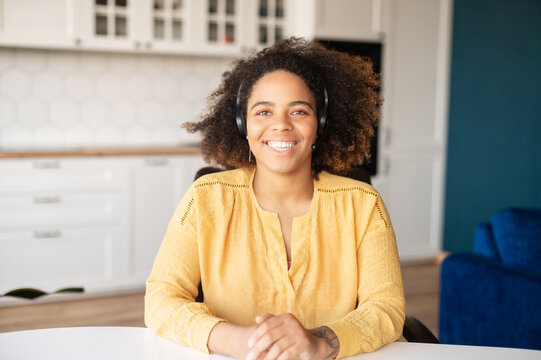 Headshot Of Friendly And Smiling African-American Woman Wearing Headset Sitting Indoor At The Home Office. Mixed-race Female With Curly Hairstyle Wearing In Yellow Shirt Looking At The Camera Friendly