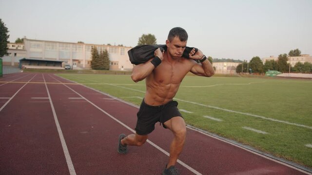 Bodybuilder in black shorts trains in the stadium and lunges with a sandbag on his shoulders