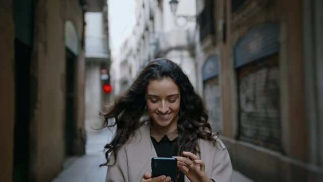 Young good-looking girl confidently walking narrow city streets and using mobile device to communicate, chat with her friends and post photos. Modern technology usability concept. Slow motion shot