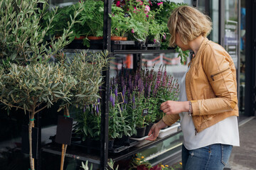 Fototapeta na wymiar Young blonde woman, owner of flower shop, corrects the plants displayed on street window. Small business. Flower trade. Floristry and gardening.Flower shop.