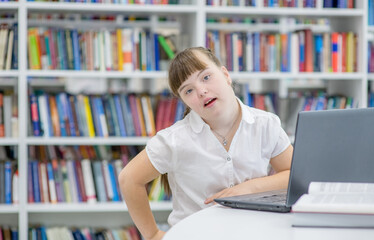 Portrait of a smiling girl with syndrome down using a laptop at library. Education for disabled...
