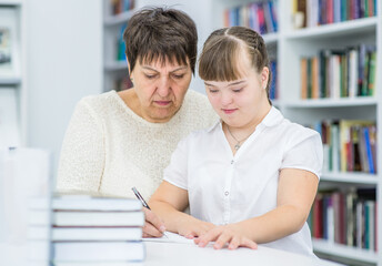 Fototapeta na wymiar Old teacher helps to girl with syndrome doing homework at library. Education for disabled children concept