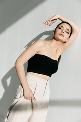 a woman in a short T-shirt and trousers holds her hand near her face and leans against the wall