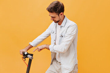 Fototapeta na wymiar Stylish bearded man in fashionable denim light clothes showing his finger on scooter and smiling on isolated orange background..