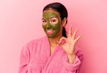 Young Venezuelan woman wearing a bathrobe and facial mask isolated on pink background cheerful and confident showing ok gesture.
